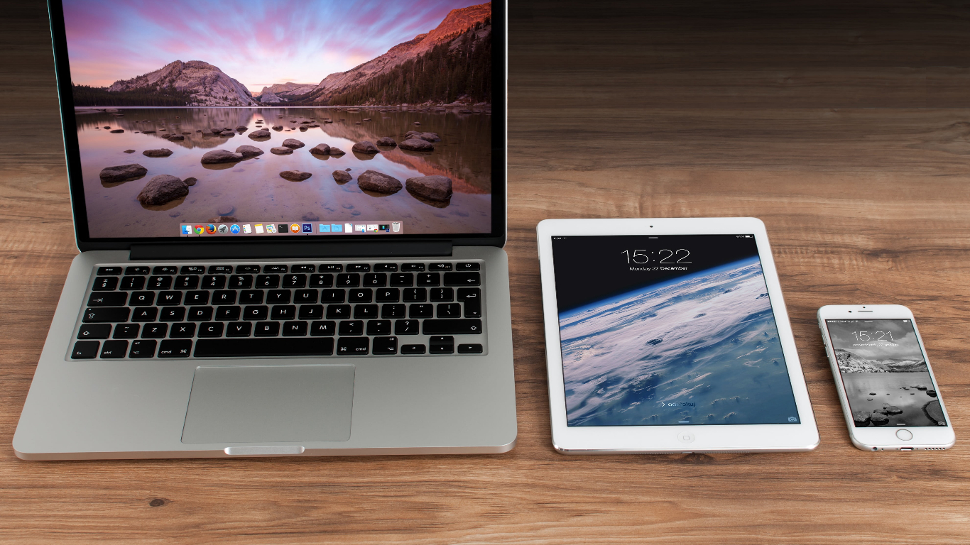 a computer, a tablet, and a phone lined up next to each other on a wooden surface