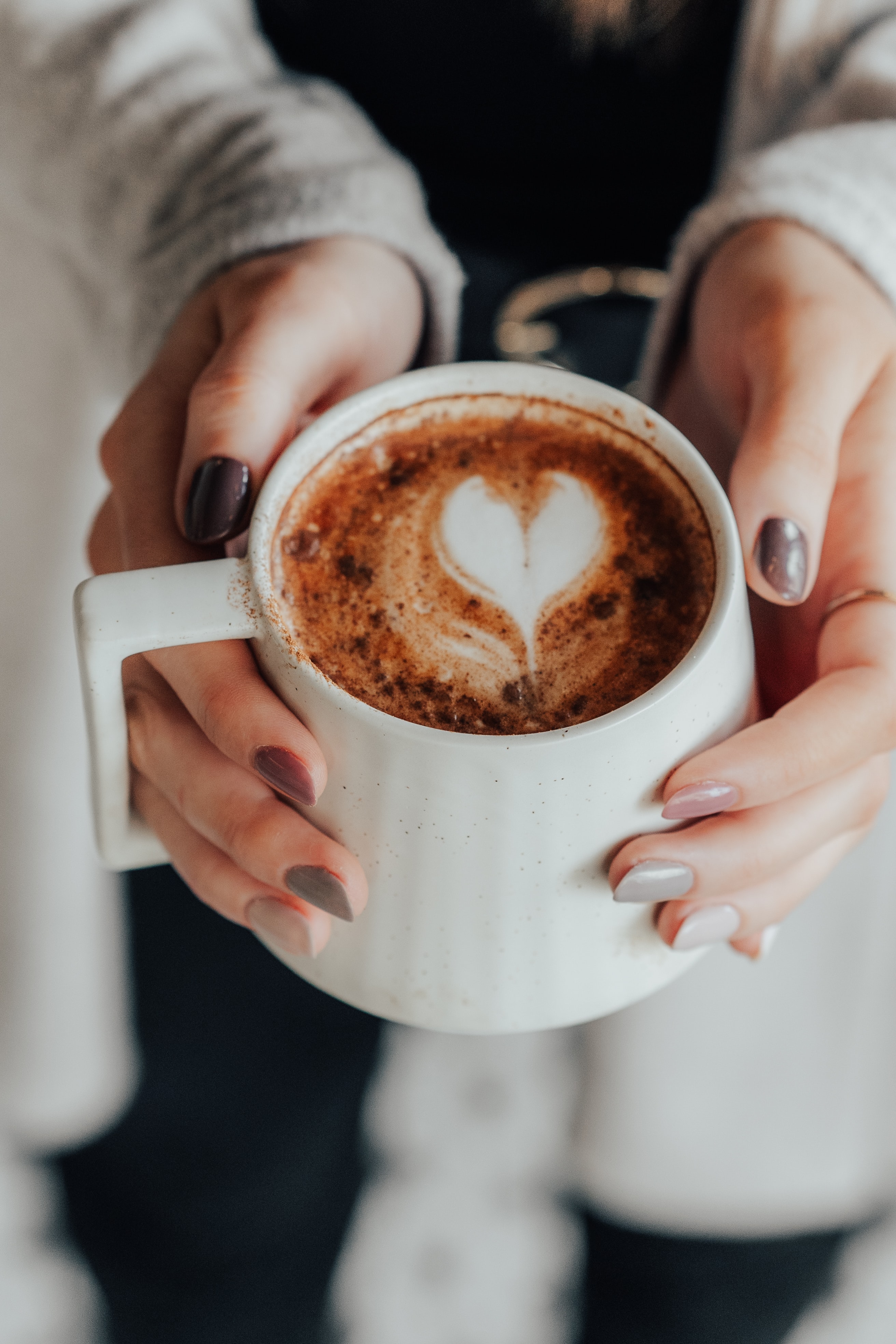 manicured hands holding a white mug with hot chocolate inside.  White milk foam is in the shape of a heart.